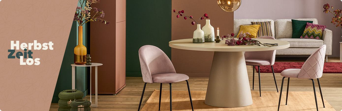 Herbstzeitlos. Esszimmer mit beigem, runden Esstisch, 2 rosa Polsterstühlen, einem grauen Sofa, Teppiche in Gelb und Dunkelrot, Kissen, Vasen und herbstliche Blumen.