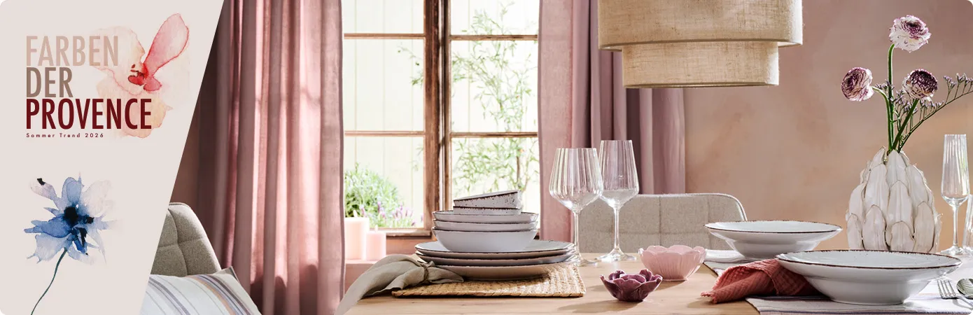 Farben der Provence - Sommertrend 2026. Gedeckter Holztisch mit Tischläufer, Platzsets, grauem Geschirr mit dunklem Rand, Weingläsern, Blumenvase und Teelichthaltern in Blütenform, Pendelleuchte mit Stoffschirm