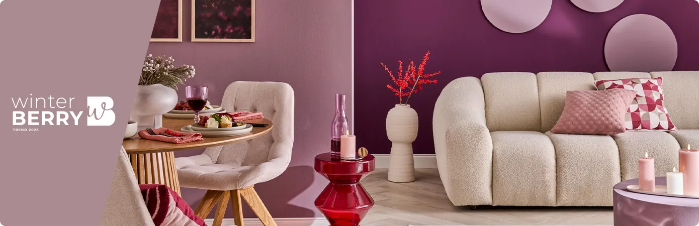 Winterberry Trend 2026. Wohnzimmer: Gedeckter Holzesstisch, cremefarbiger Polsterstuhl, roter Glastisch mit Deko, cremefarbige Bodenvase, cremefarbiges Sofa mit rosa und mehrfarbig gemustertem Kissen, rosa Couchtisch mit Deko. 